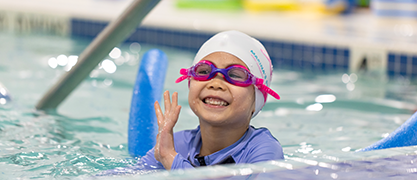 "Smiling young swimmer with goggles and a swim cap using a noodle during a lesson at Marina Swim School."