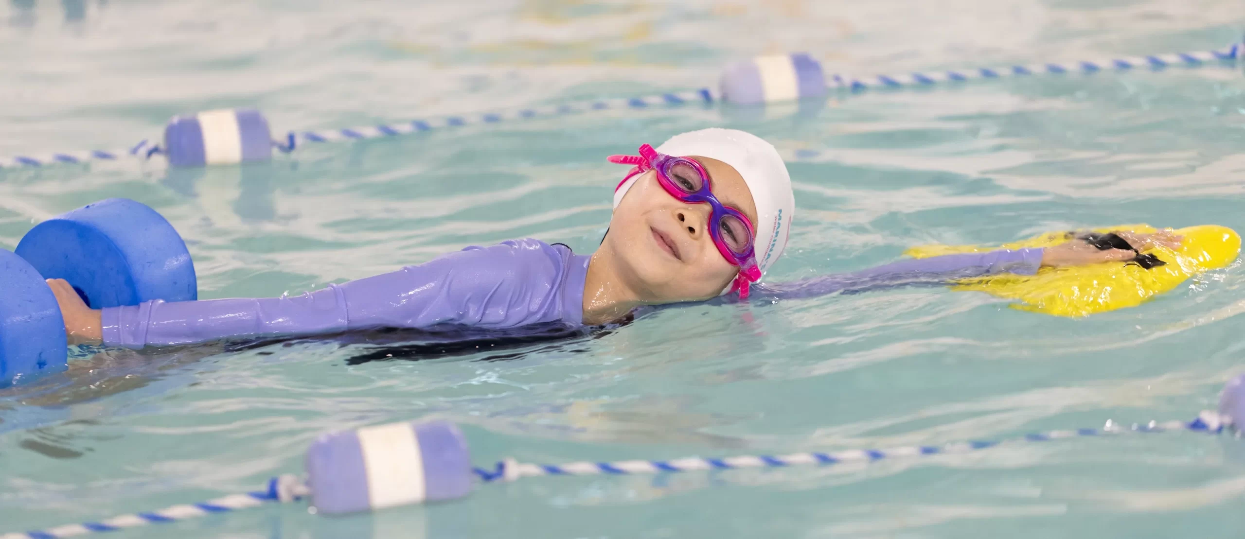 Young swimmer at Marina’s Swim School swim camp using a kickboard and pool weights during a swimming lesson.