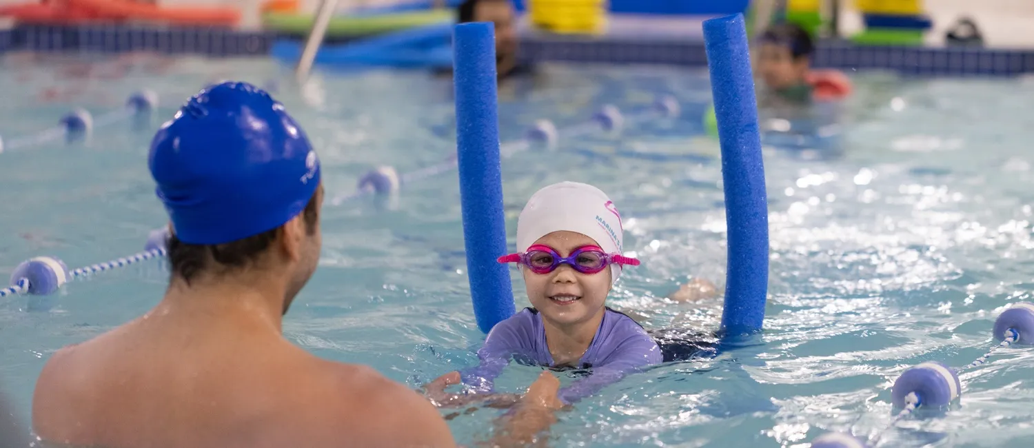 Marina’s Swim School instructor teaching a young swimmer with goggles and a swim cap using pool noodles during a private lesson.