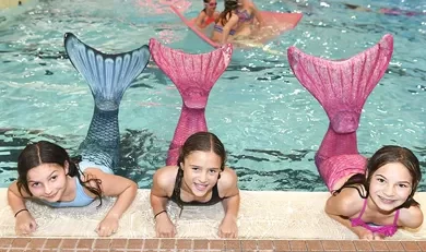 Kids in Marina’s Swim School Mermaid Class wearing colorful mermaid tails and learning swimming skills in a warm indoor pool.