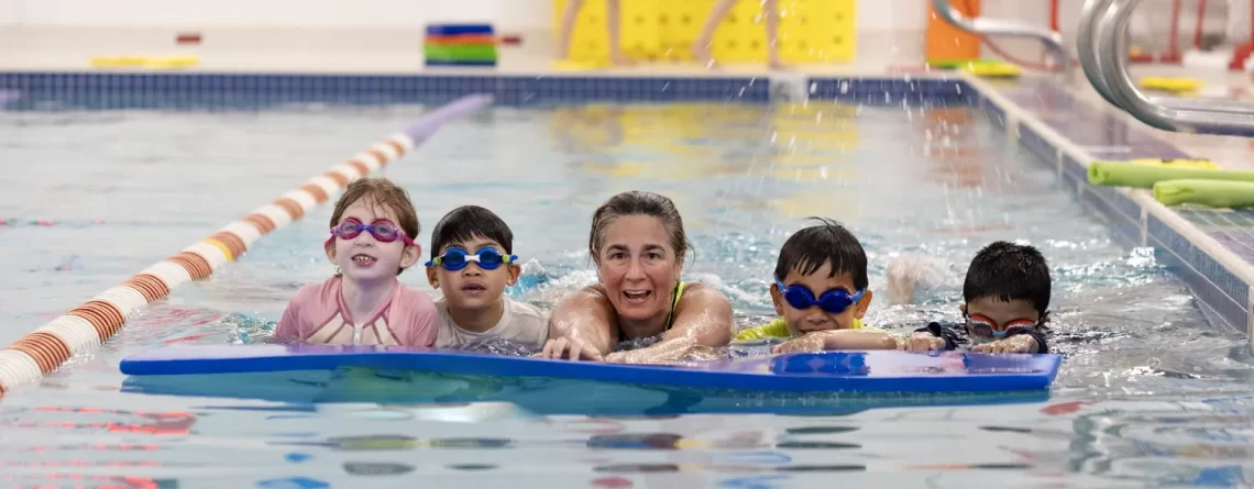 Marina’s Swim School instructor leading a group of young swimmers practicing with a large kickboard in a warm indoor pool.